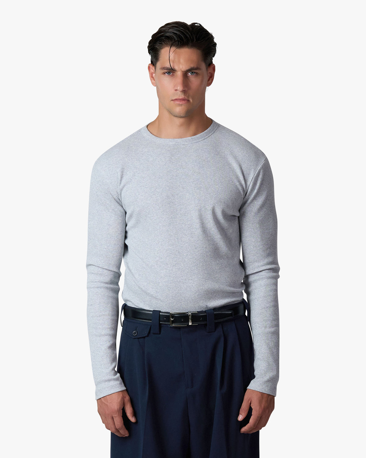 A man wearing a light gray long-sleeve crewneck shirt tucked into dark navy trousers with a black belt, photographed against a clean white studio background.
